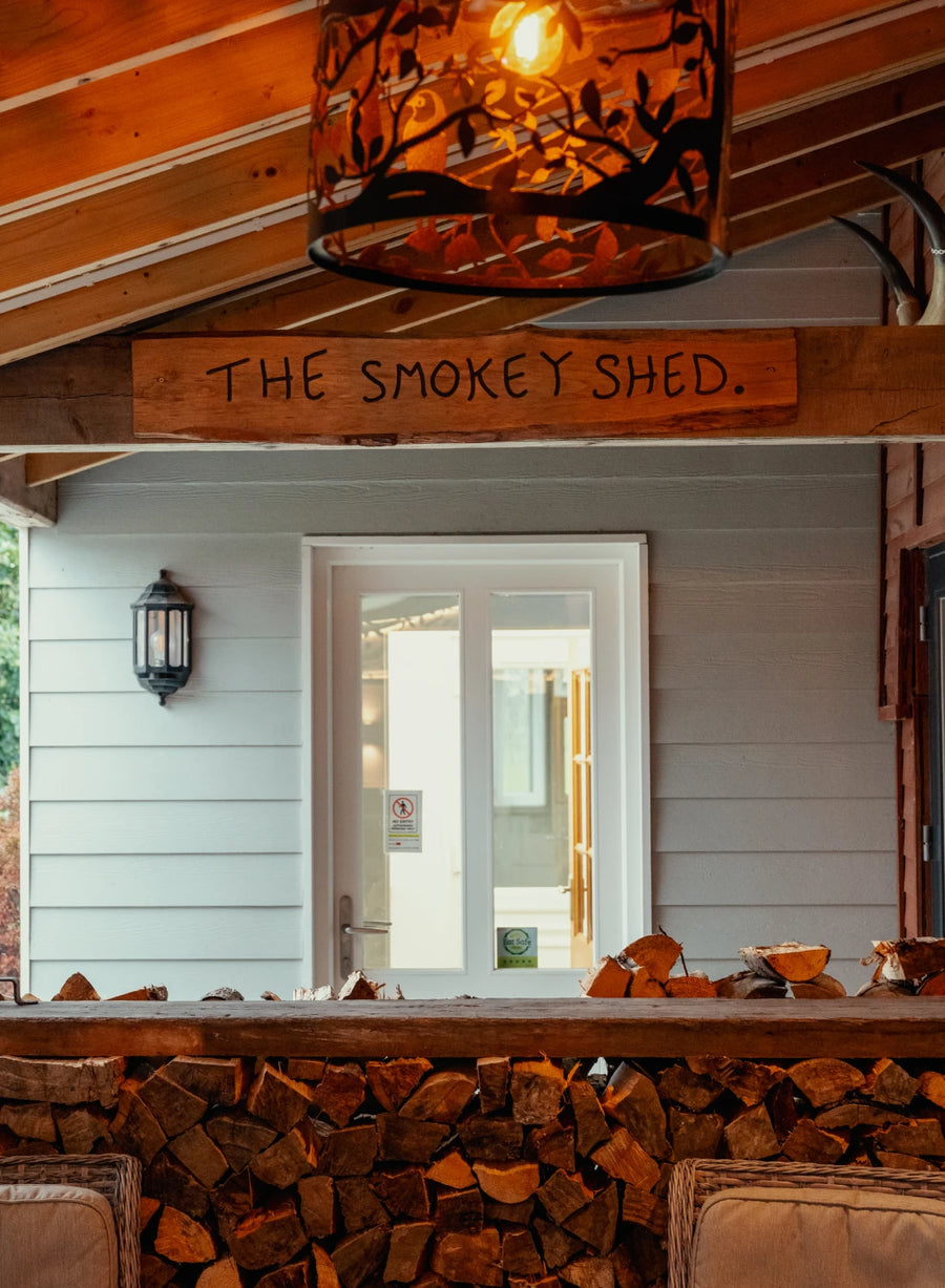 The Smokey Shed sign above the door, with oak logs stacked outside